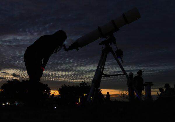 A person is silhouetted against a sunset, looking through a telescope. The sky is filled with dark clouds, and other people and trees are faintly visible in the background.