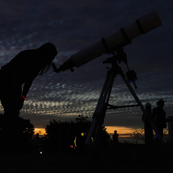 A person is silhouetted against a sunset, looking through a telescope. The sky is filled with dark clouds, and other people and trees are faintly visible in the background.